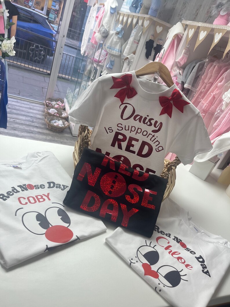 May include: White and black t-shirts with Red Nose Day graphics. One shirt has the text "Daisy is Supporting RED NOSE DAY" with red bows. Other shirts feature cartoon faces and names like "COBY" and "Chloe".