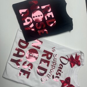 May include: Two t-shirts, one black and one white, with the words "RED NOSE DAY" in a glittery red font. The white shirt has red bows and the text "Daisy is supporting RED NOSE DAY".