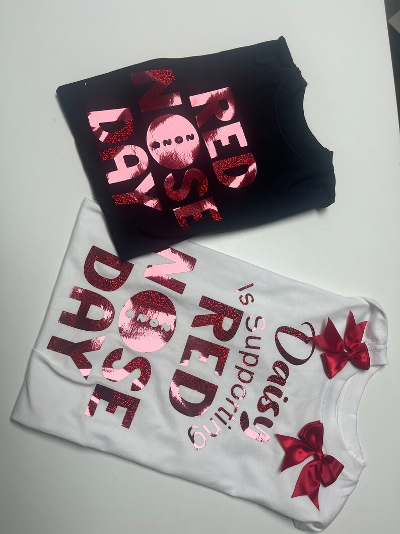 May include: Two t-shirts, one black and one white, with the words "RED NOSE DAY" in a glittery red font. The white shirt has red bows and the text "Daisy is supporting RED NOSE DAY".