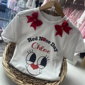 May include: White t-shirt with red bows on the shoulders. The shirt features a cartoon face design with a red heart nose and the text "Red Nose Day Chloe". The shirt is displayed in a woven basket.