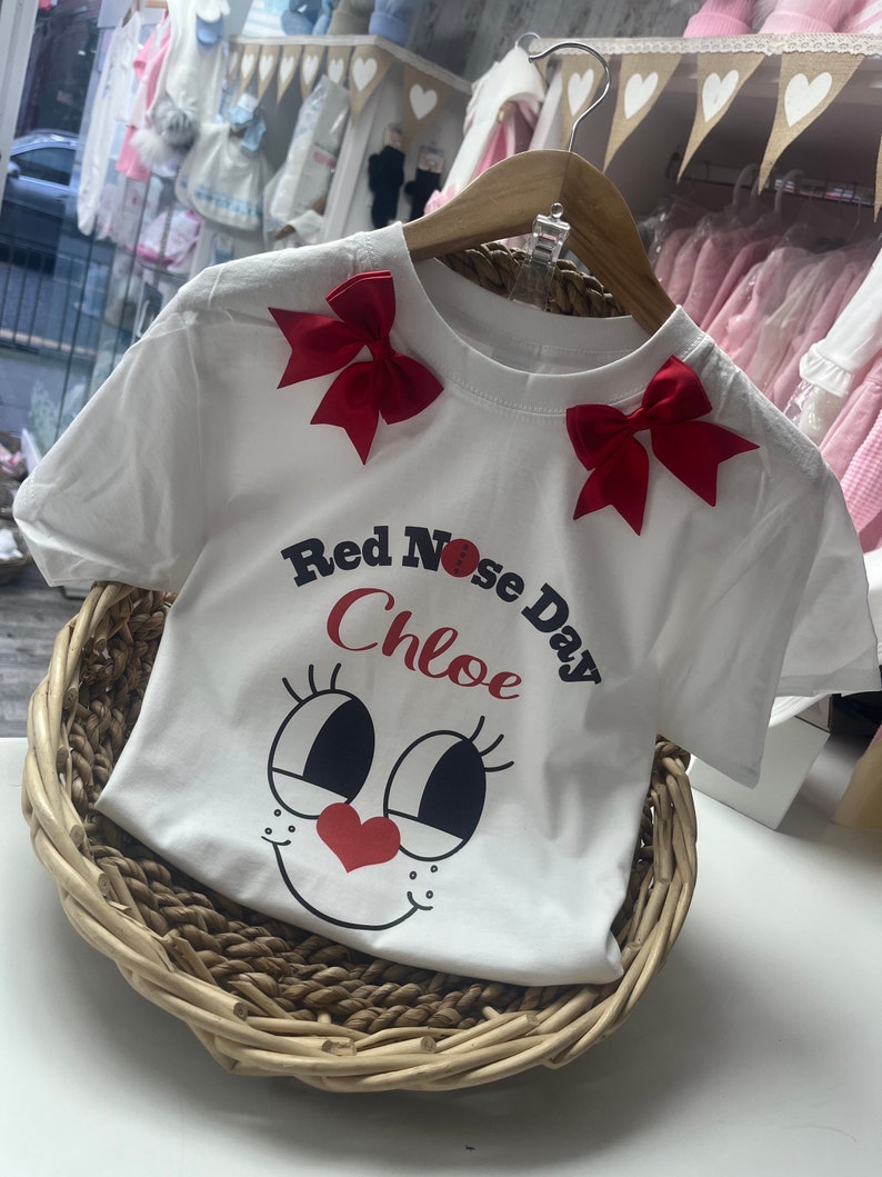 May include: White t-shirt with red bows on the shoulders. The shirt features a cartoon face design with a red heart nose and the text "Red Nose Day Chloe". The shirt is displayed in a woven basket.
