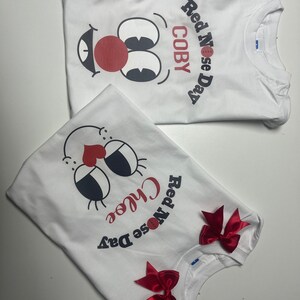 May include: Two white t-shirts with a cartoon face design featuring a red nose, black eyes, and a smiling mouth. One shirt says "COBY" and "Red Nose Day." The other says "Chloe" and "Red Nose Day" with red bows.