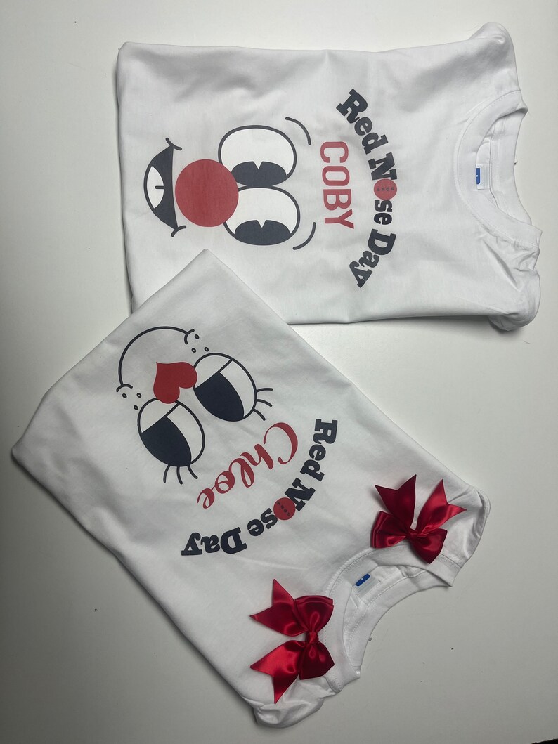 May include: Two white t-shirts with a cartoon face design featuring a red nose, black eyes, and a smiling mouth. One shirt says "COBY" and "Red Nose Day." The other says "Chloe" and "Red Nose Day" with red bows.