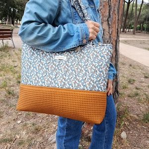 Può includere: Una grande borsa tote con un motivo floreale blu, bianco e marrone e un fondo arancione-marrone strutturato. La borsa ha una tracolla abbinata e una piccola etichetta. La borsa viene portata all'aperto.