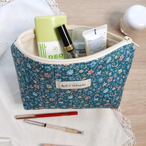 Può includere: Una pochette trapuntata color verde acqua con un piccolo motivo floreale e una cerniera color crema. Un'etichetta rettangolare in tessuto con la scritta "ArtiTextures" è centrata sul davanti. La pochette è riempita con vari prodotti di cura.
