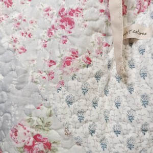 May include: A quilted fabric featuring a patchwork design with floral patterns in shades of pink, white, and light blue. The textile has a visible stitched texture and includes a small beige fabric tag with the text "Petit textures" attached.