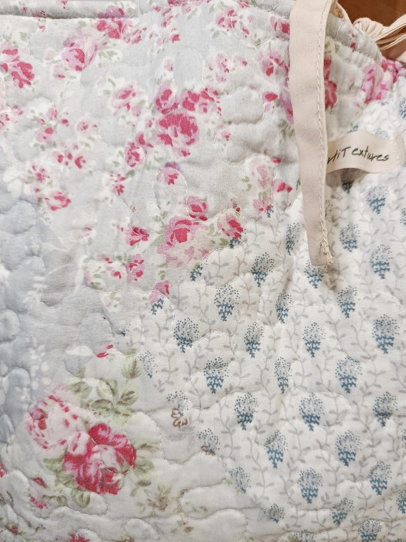 May include: A quilted fabric featuring a patchwork design with floral patterns in shades of pink, white, and light blue. The textile has a visible stitched texture and includes a small beige fabric tag with the text "Petit textures" attached.
