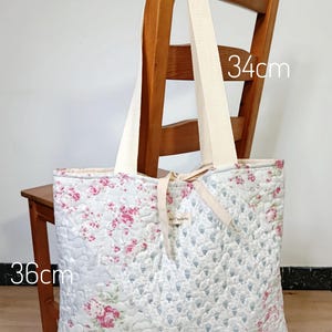 May include: A quilted tote bag featuring a floral pattern with pink roses and blue sprigs on a light background. The bag has cream-colored fabric handles and a small central label. Dimensions are 36cm by 44cm by 8cm, with a 34cm handle drop.