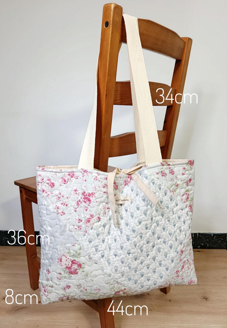 May include: A quilted tote bag featuring a floral pattern with pink roses and blue sprigs on a light background. The bag has cream-colored fabric handles and a small central label. Dimensions are 36cm by 44cm by 8cm, with a 34cm handle drop.
