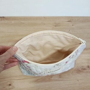 May include: A quilted fabric pouch with a zipper closure, featuring a floral pattern in shades of pink and green on a white background. The interior is lined with plain cream fabric, and a small white label with the text ArtiTextures is attached.