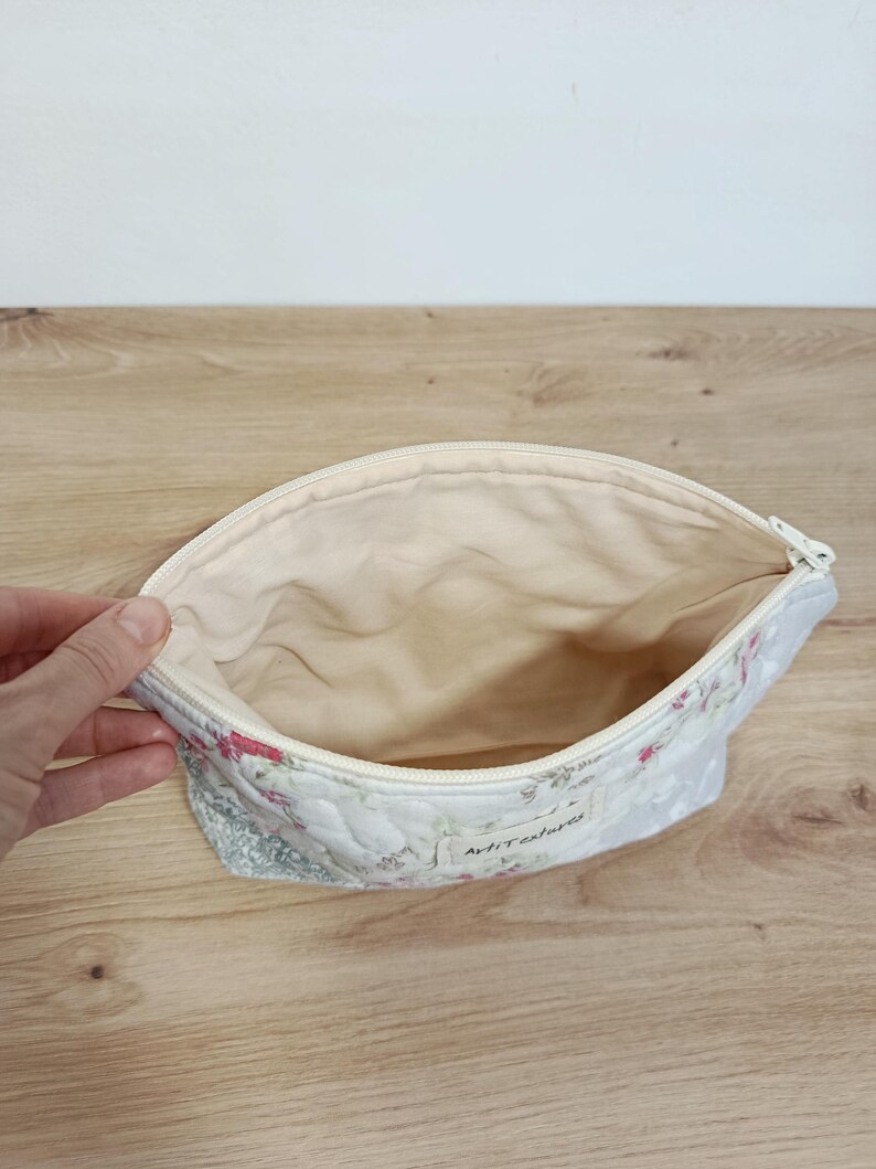 May include: A quilted fabric pouch with a zipper closure, featuring a floral pattern in shades of pink and green on a white background. The interior is lined with plain cream fabric, and a small white label with the text ArtiTextures is attached.