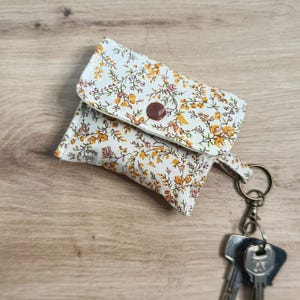 Puede incluir: Pequeño monedero con estampado floral, con flores naranjas y amarillas sobre fondo blanco. Cierre con botón marrón. Llavero y llaves adjuntas. La tela es floreada.