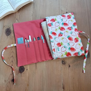 Könnte beinhalten: Ein korallenfarbenes Stifte- und Bleistift-Etui mit einem Buchumschlag mit Erdbeermuster. Die Rolle ist geöffnet und zeigt Stifte und Bleistifte in einzelnen Fächern. Im Hintergrund ist ein Buch zu sehen, und die Rolle ist mit einem passenden Band mit Erdbeermuster gebunden.