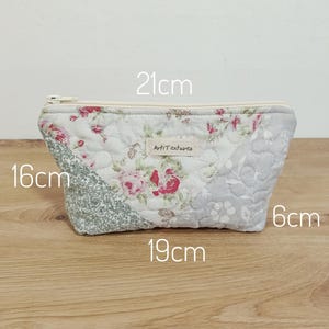 May include: A quilted fabric pouch featuring a floral pattern in shades of pink, green, and grey. A white zipper runs along the top. A small label reads "ArtiTextures". Dimensions are printed as 21cm wide, 16cm high, 19cm base width, and 6cm deep.