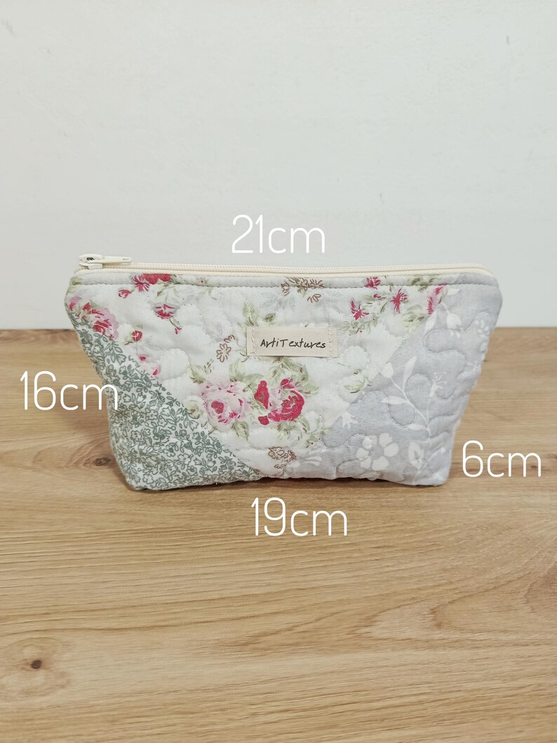 May include: A quilted fabric pouch featuring a floral pattern in shades of pink, green, and grey. A white zipper runs along the top. A small label reads "ArtiTextures". Dimensions are printed as 21cm wide, 16cm high, 19cm base width, and 6cm deep.