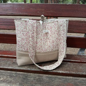 Può includere: Una borsa tote trapuntata con un motivo floreale rosa e bianco, e un fondo beige a trama waffle. La borsa ha un manico abbinato e una piccola etichetta con la scritta "Apt Tales". La borsa è appoggiata su una panchina di legno.