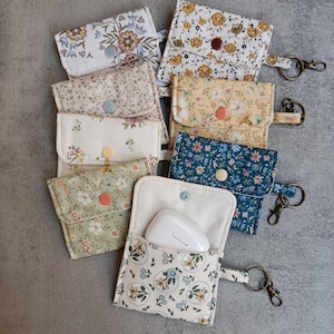 Può includere: Una collezione di pochette in tessuto con motivi floreali in vari colori, tra cui blu, giallo e verde. Ogni pochette ha una chiusura a bottone e un piccolo portachiavi. Una pochette è aperta, rivelando un dispositivo elettronico bianco.