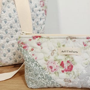 May include: A quilted fabric pouch featuring a floral pattern with pink roses and green foliage, a cream-colored zipper, and a rectangular fabric label reading "ArtiTextures". A matching quilted bag with a cream strap is visible in the background.