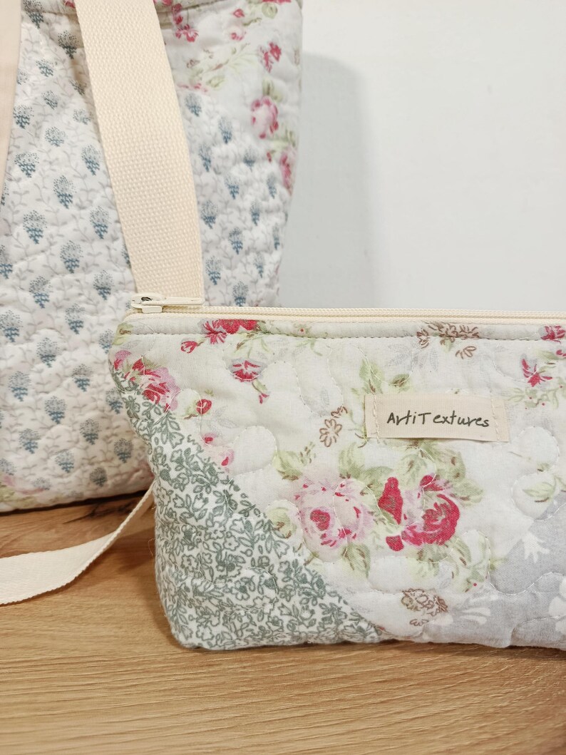 May include: A quilted fabric pouch featuring a floral pattern with pink roses and green foliage, a cream-colored zipper, and a rectangular fabric label reading "ArtiTextures". A matching quilted bag with a cream strap is visible in the background.