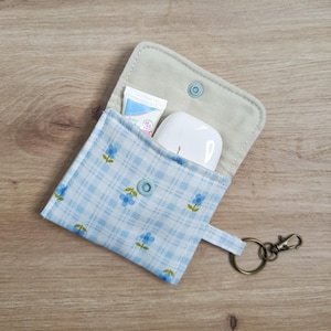 May include: Open, light blue floral patterned pouch with a snap closure, holding a small tube of lotion and a white wireless earbud case. The pouch has a key ring attached.