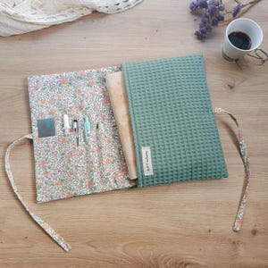 Op de afbeelding: Een saliegroene stoffen hoes met wafelstructuur en een bloemige voering met roze en oranje bloesems. De hoes bevat meerdere vakjes voor pennen, een beige label met de tekst Artitcatures en twee lange stoffen linten om te strikken.