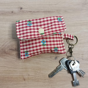 Può includere: Un piccolo astuccio in tessuto a quadretti rossi e bianchi con chiusura a bottone, ornato da dettagli floreali blu. Attaccato a un portachiavi con diverse chiavi. L'astuccio è progettato per contenere piccoli oggetti.