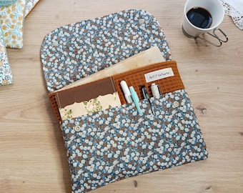 May include: A fabric document organizer featuring a blue and white floral print exterior with a waffle-textured brown interior. It includes a flap closure with metal snaps, internal pockets holding pens and notebooks, and a label reading ArtiTectures.
