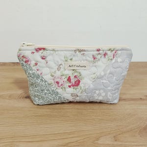 May include: A quilted zippered pouch featuring a patchwork design of floral patterns in shades of pink, green, and grey. A small rectangular fabric label with the text ArtiTextures is sewn onto the front center of the quilted fabric.