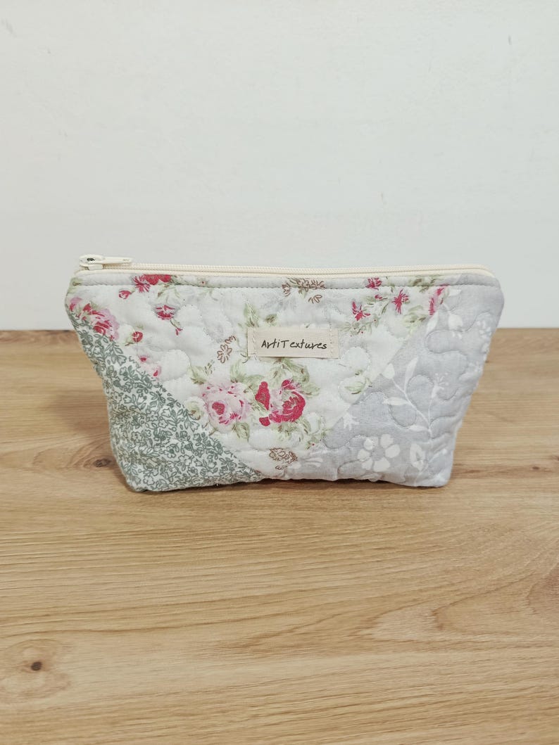May include: A quilted zippered pouch featuring a patchwork design of floral patterns in shades of pink, green, and grey. A small rectangular fabric label with the text ArtiTextures is sewn onto the front center of the quilted fabric.