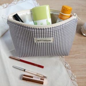May include: A grey and white gingham fabric pouch with a zipper closure and a central label reading ArtiTextures. The open pouch contains a light green bottle, an orange bottle, and a black tube. Three cosmetic pencils lie in front of the pouch.
