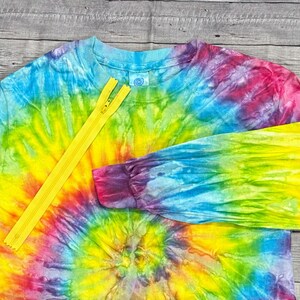 May include: A long-sleeved tie-dye shirt in a vibrant spiral pattern of rainbow colors. A bright yellow zipper is placed diagonally across the shirt. The shirt has a crew neck and cuffed sleeves.