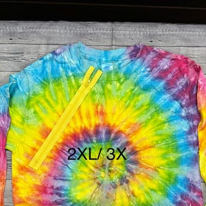 May include: A long-sleeved tie-dye shirt in a spiral pattern of rainbow colors, with a yellow zipper. The shirt is labeled "2XL/3X". The shirt is on a wooden surface.