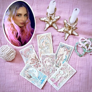 May include: A collection of tarot cards with illustrations in blue, purple, and gold, next to a portrait of a woman, two star-shaped candle holders with white candles, a white seashell, and two bracelets. The items are on a pink surface.