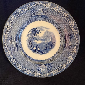 Puede incluir: Plato decorativo azul y blanco con una escena de paisaje en el centro. El plato presenta un borde detallado con un patrón de celosía y diseño de vid. El plato redondo tiene una estética clásica y vintage.