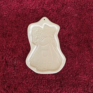 Puede incluir: Cortador de galletas de cerámica blanquecina con forma de mujer sosteniendo una cesta. El cortador tiene un borde festoneado y un agujero para colgarlo. La mujer lleva un vestido con delantal y un gorro.
