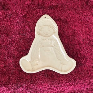 Puede incluir: Adorno de pared de cerámica blanquecina con forma de niño sentado. El niño lleva un gorro y un peto, con una cara sonriente. Tiene un agujero en la parte superior para colgar.