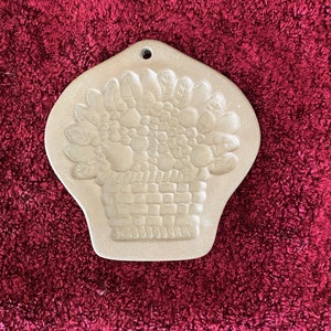 Puede incluir: Adorno de pared de cerámica beige con una cesta llena de fruta. La cesta tiene un diseño de ladrillo, y la fruta está detallada con hojas y formas redondas. Hay un pequeño agujero en la parte superior para colgar.