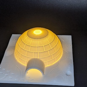 May include: A 3D-printed igloo night light. The white structure has a domed roof and an arched entrance, with a warm yellow light emanating from within. The igloo sits on a white base with textured details, creating a cozy, illuminated scene.