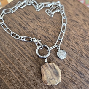 May include: Silver-toned chain bracelet with a large, irregular-shaped wooden pendant. The bracelet features a lobster clasp and a small circular charm with a tree design. The wooden pendant has a natural, light brown and tan wood grain.