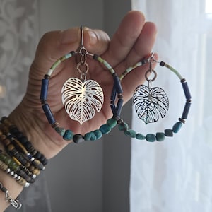 May include: A pair of hoop earrings featuring silver-toned monstera leaf charms suspended from copper-colored rings. The hoops are decorated with a mix of cylindrical dark blue beads, small turquoise-colored stones, and light green accents.