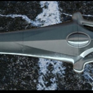 May include: A silver-colored sword with a black handle and a detailed design. The sword is lying on a dark, textured surface.