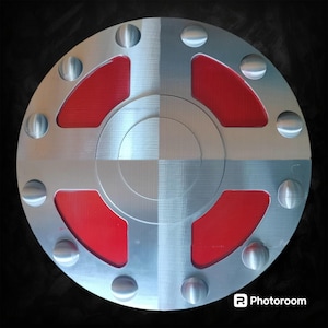 May include: A circular metal object with a brushed silver finish. It features a series of evenly spaced, raised, rounded rivets around the perimeter. The center has a cross-shaped design with red inserts, creating a striking contrast against the silver.