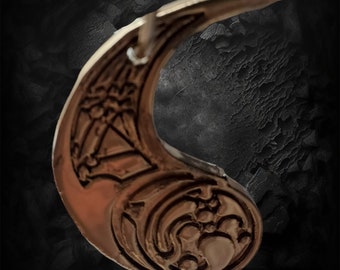 May include: A silver-colored metal pendant shaped like a crescent moon with intricate designs. The pendant has a textured surface and a smooth, rounded edge.
