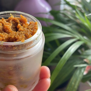 May include: A clear glass jar filled with a thick, orange-brown scrub. The scrub has a grainy texture and is likely a body or face scrub.