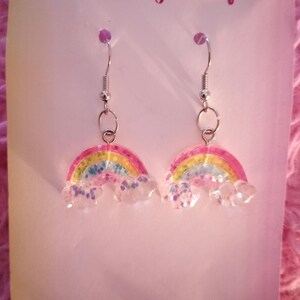 May include: A pair of handmade rainbow earrings with silver hooks. The earrings feature a colorful rainbow design with pink, yellow, blue, and green hues, and clear cloud accents. The text "Just A Girly Boutique" and "Handmade Jewelry" are visible.