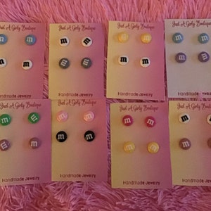 Puede incluir: Un conjunto de pendientes de botón hechos a mano con diseños de caramelos M&M's de colores. Cada par se muestra en una tarjeta pequeña con el texto "Just A Girly Boutique" y "Handmade Jewelry".