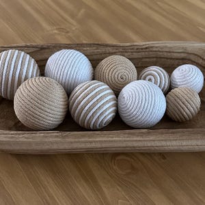 K&ouml;nnte beinhalten: Eine Holzschale gef&uuml;llt mit neun dekorativen Kugeln. Die Kugeln bestehen aus einem gewebten Material in Beige- und Wei&szlig;t&ouml;nen. Einige Kugeln haben ein Streifenmuster, andere sind einfarbig oder spiralf&ouml;rmig.