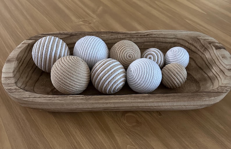 K&ouml;nnte beinhalten: Eine Holzschale gef&uuml;llt mit neun dekorativen Kugeln. Die Kugeln bestehen aus einem gewebten Material in Beige- und Wei&szlig;t&ouml;nen. Einige Kugeln haben ein Streifenmuster, andere sind einfarbig oder spiralf&ouml;rmig.