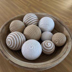 K&ouml;nnte beinhalten: Eine Holzschale gef&uuml;llt mit dekorativen Kugeln. Die Kugeln bestehen aus gewebtem Seil in Beige, Wei&szlig; und Creme. Einige Kugeln haben Streifenmuster, w&auml;hrend andere einfarbig sind. Die Schale ist aus hellbraunem Holz. Die Kugeln haben einen Durchmesser von ca. 5-8 cm.