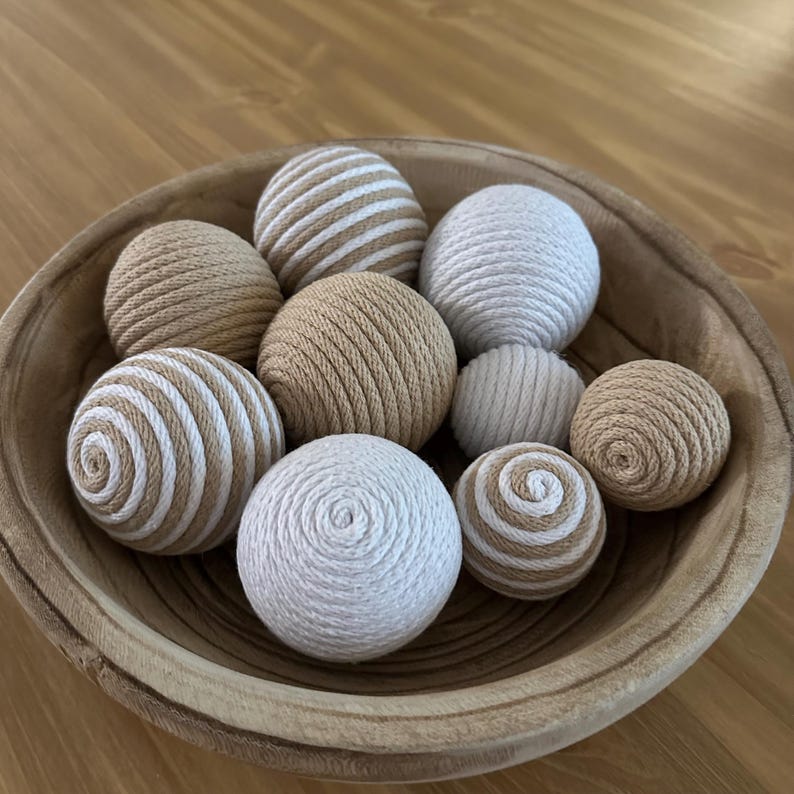 K&ouml;nnte beinhalten: Eine Holzschale gef&uuml;llt mit dekorativen Kugeln. Die Kugeln bestehen aus gewebtem Seil in Beige, Wei&szlig; und Creme. Einige Kugeln haben Streifenmuster, w&auml;hrend andere einfarbig sind. Die Schale ist aus hellbraunem Holz. Die Kugeln haben einen Durchmesser von ca. 5-8 cm.